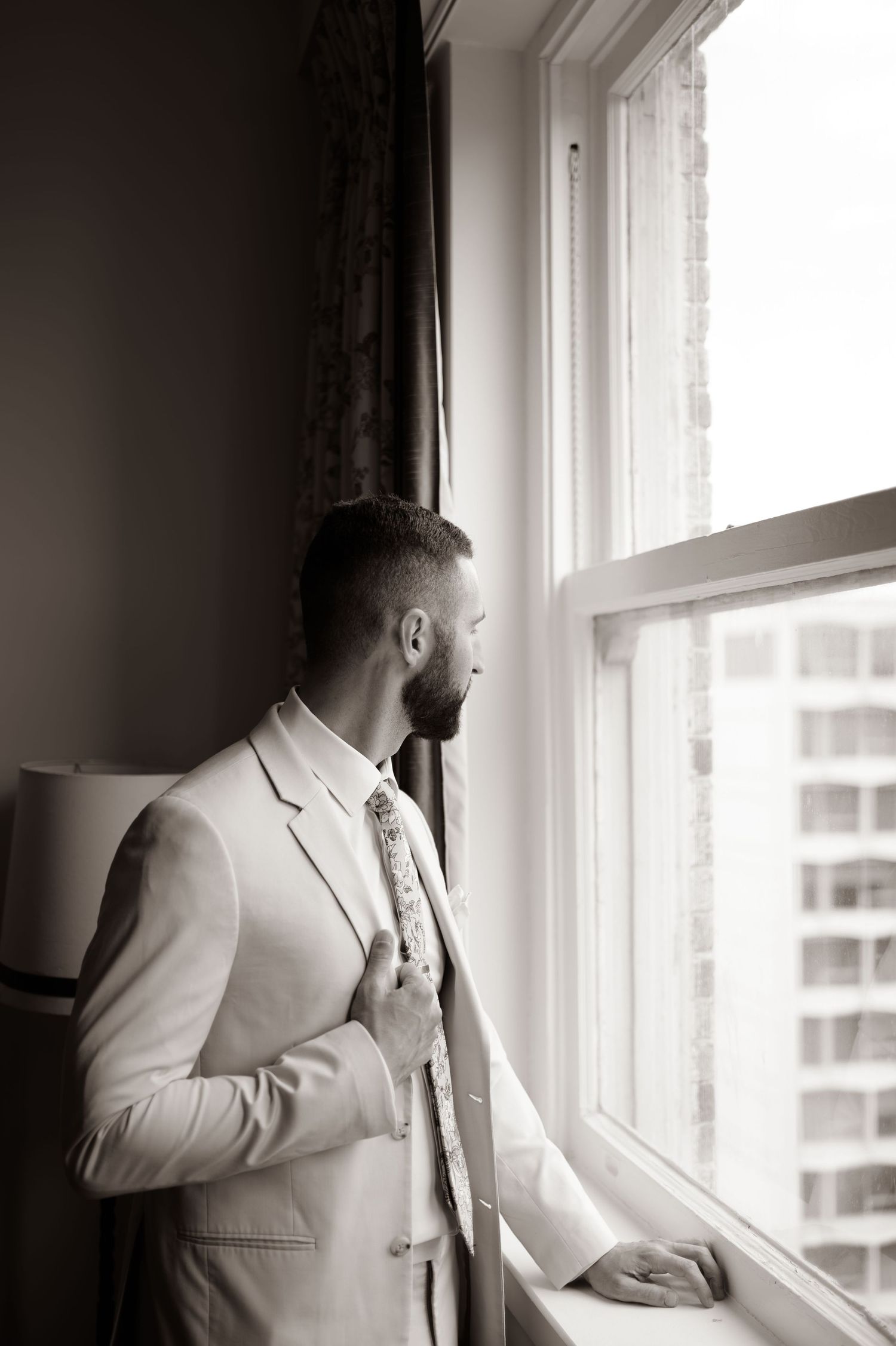 Person in light suit gazes thoughtfully out window in black and white portrait series.