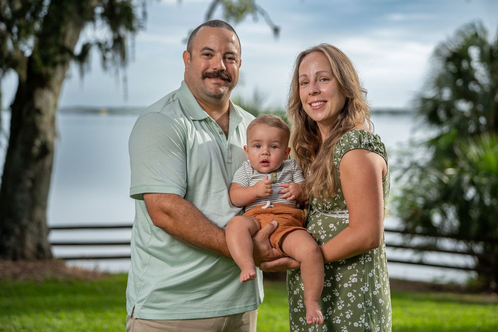 Happy family standing together near lake with trees and wooden fence, outdoor family photography in Tampa, Florida.