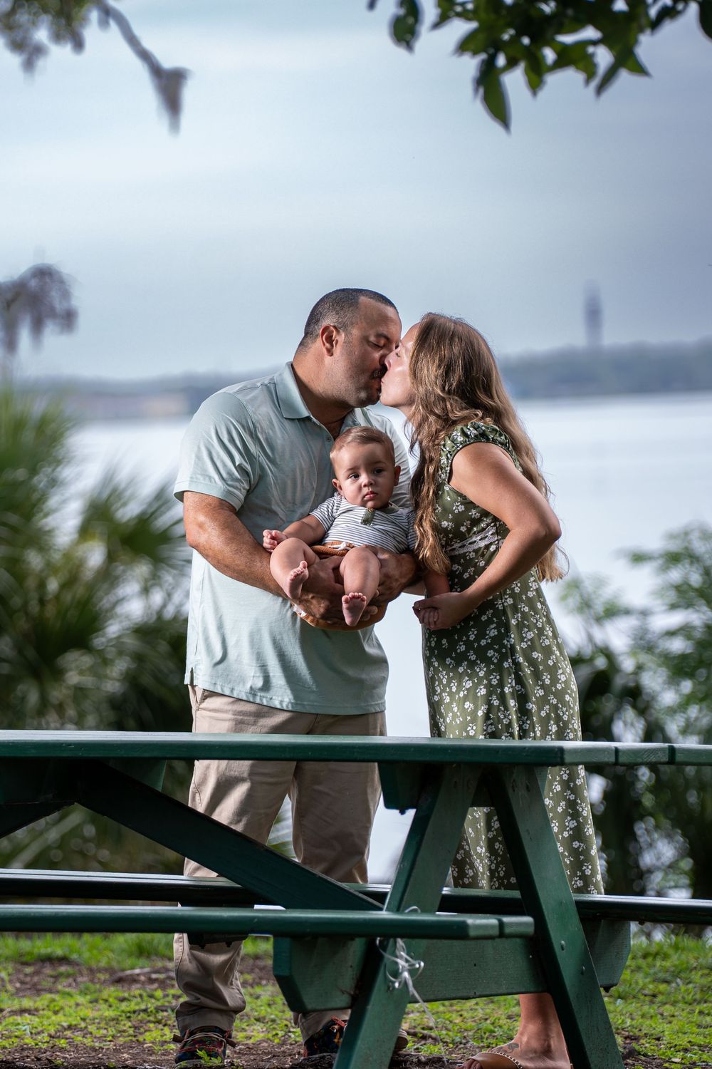 Couple with baby enjoying family time at picnic table by lake, candid outdoor lifestyle photography in Tampa, Florida.