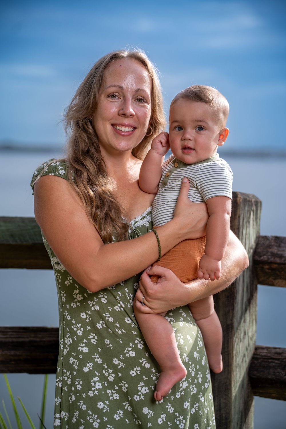 Mother in floral green dress holding baby in striped onesie by the water, outdoor family photoshoot in Tampa, Florida.