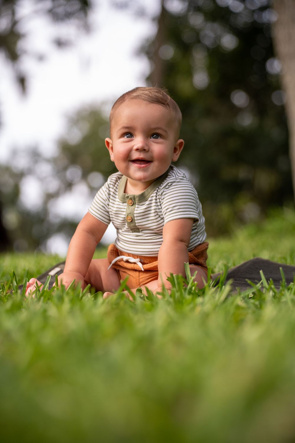 Smiling baby in striped shirt and brown pants sitting on grass, candid outdoor child portrait in Tampa, Florida.