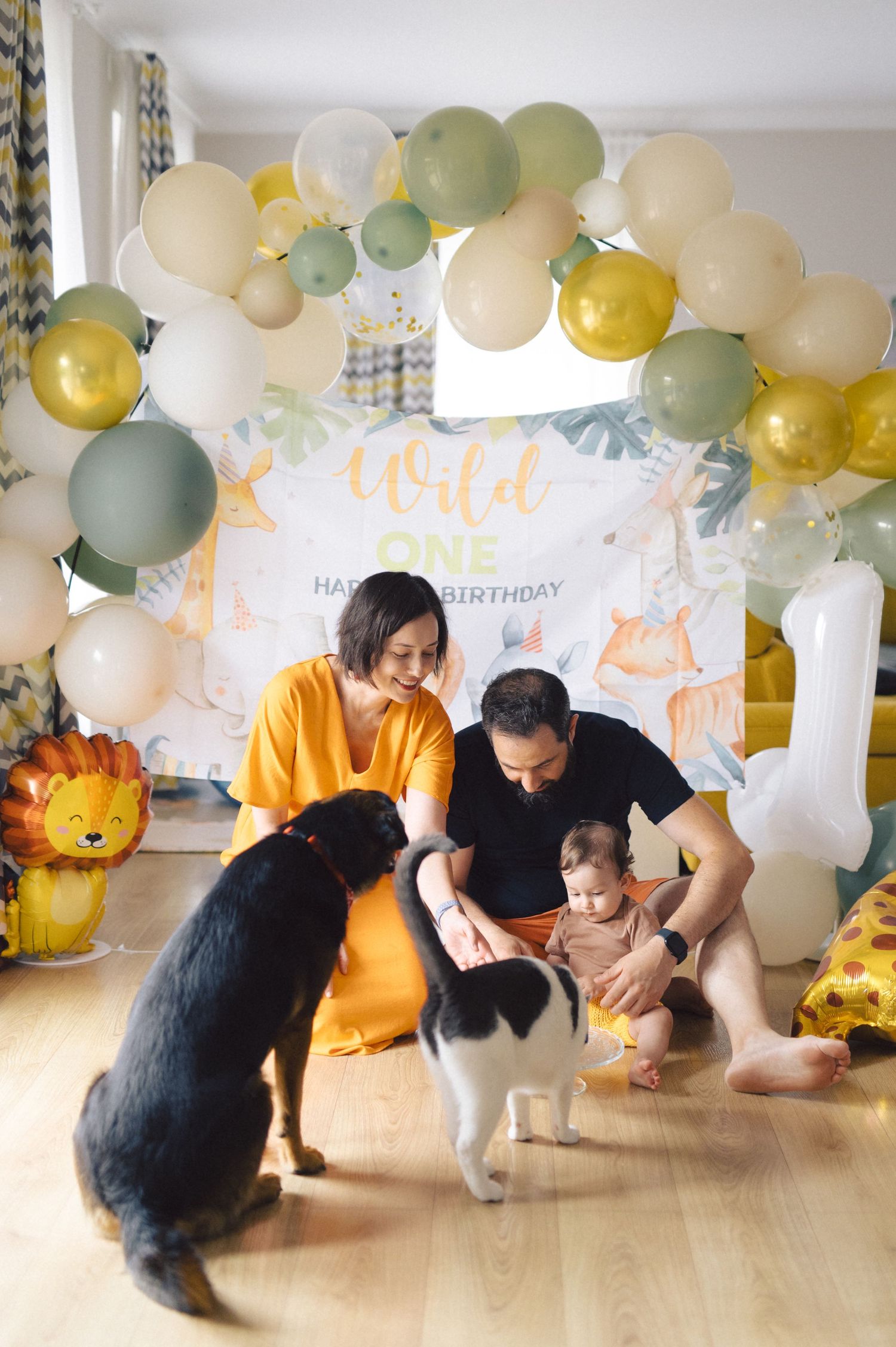 A festive celebration scene with balloons, decorations, pets and gathering under a balloon arch and banner.