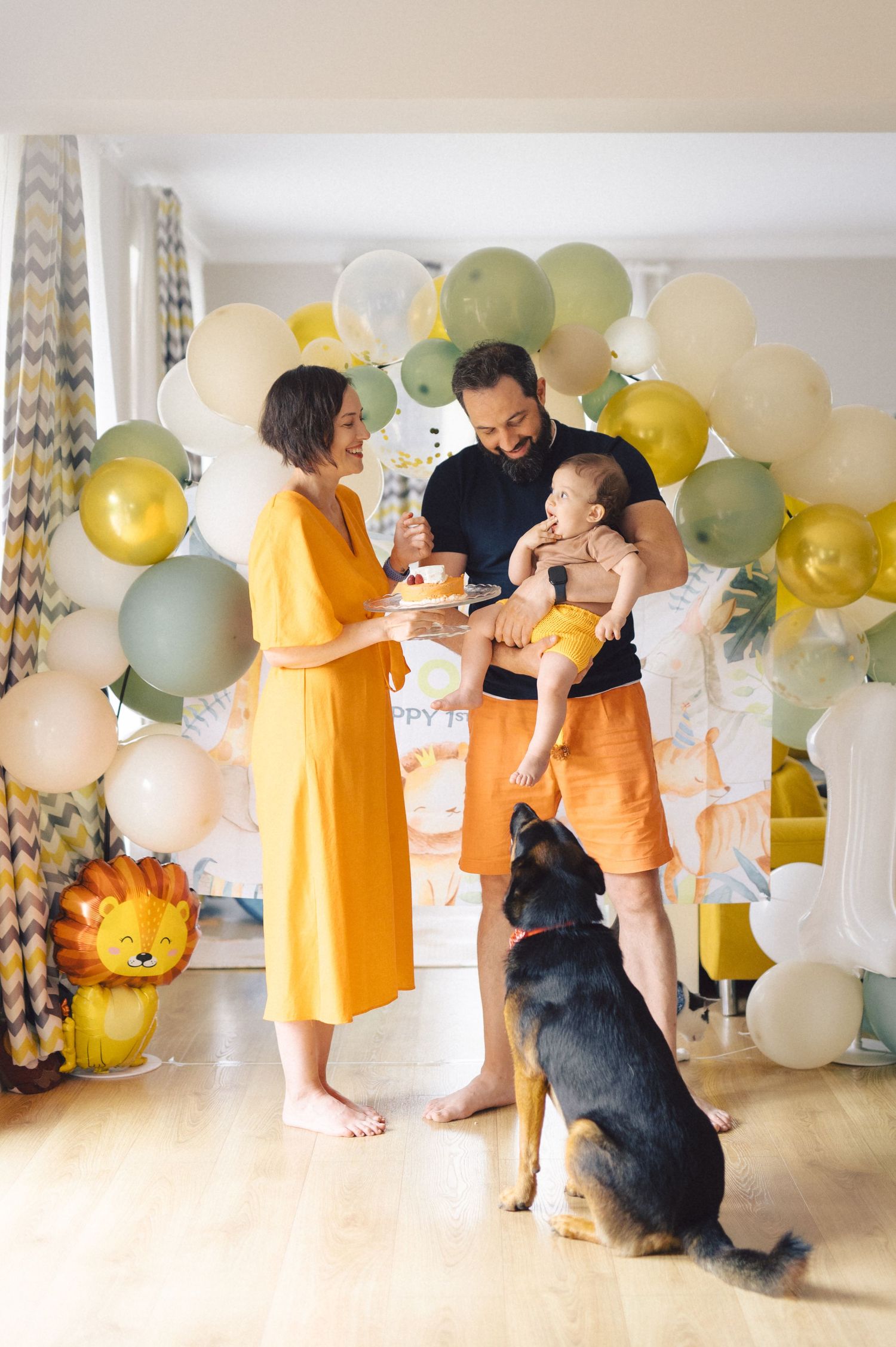 Family celebrating with balloons and their dog in front of a festive balloon arch decorated in yellow and sage colors.