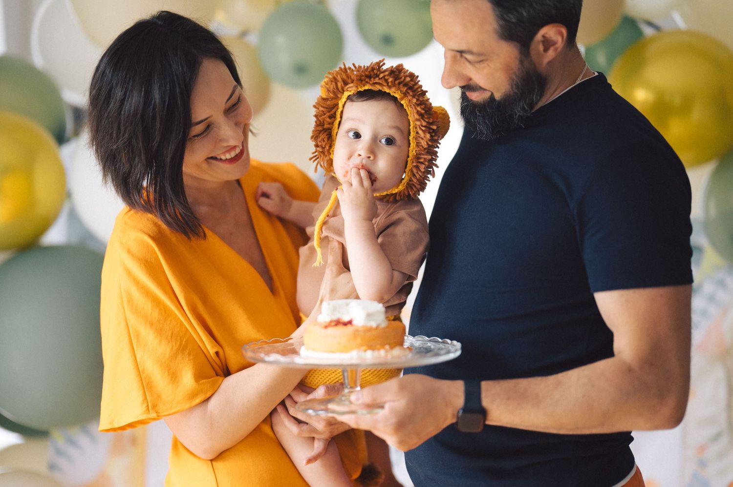 A family celebrates at a birthday party with cake and colorful balloons in the background.