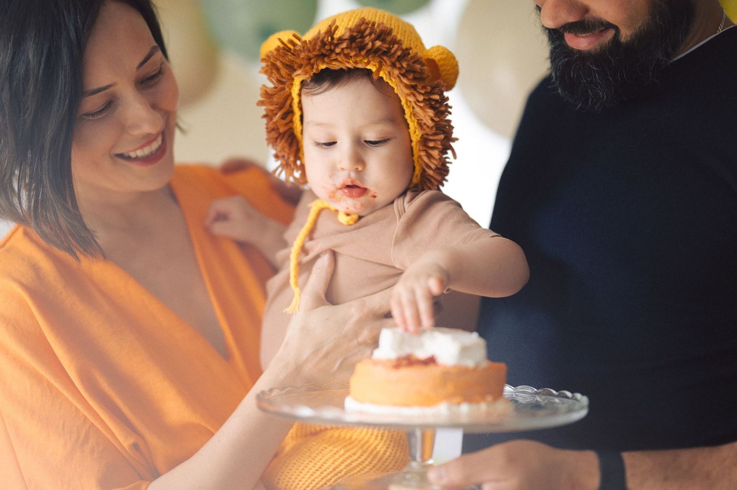 A small child wearing a lion-like costume reaches for a dessert plate while held by adults in a warm indoor setting.