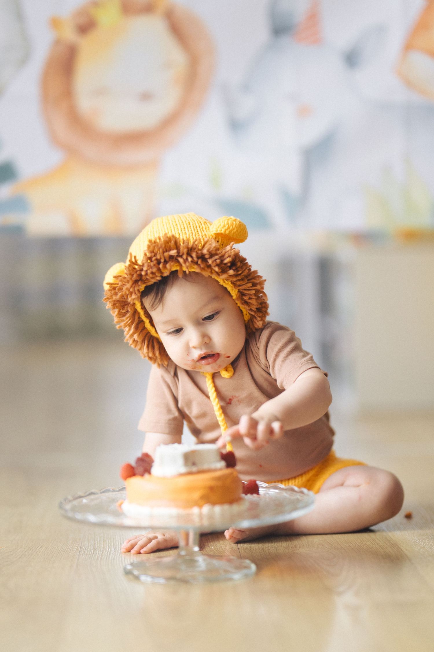 A small figure in a brown bodysuit and lion costume hat reaches for something on a glass cake stand on the floor.