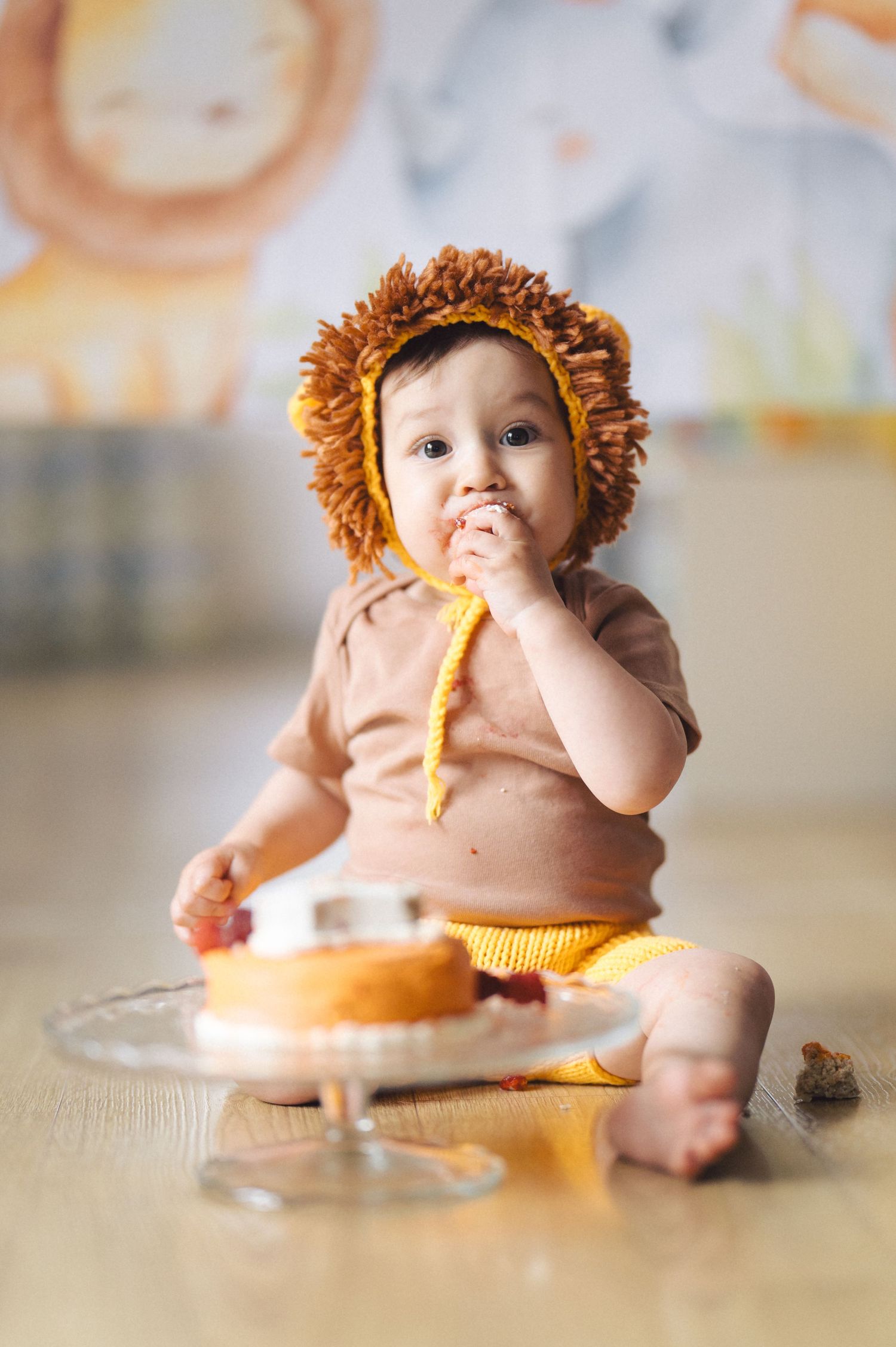 Small baby wearing a lion costume and tan shirt sits on floor eating cake from glass pedestal stand.