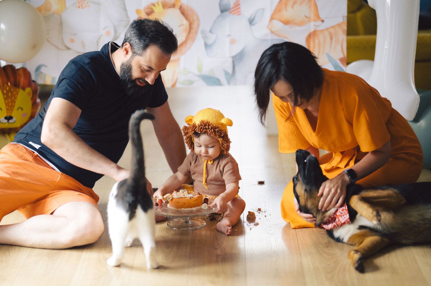 Two adults sitting on floor with a toddler wearing a fall-colored headpiece while two dogs interact with them.