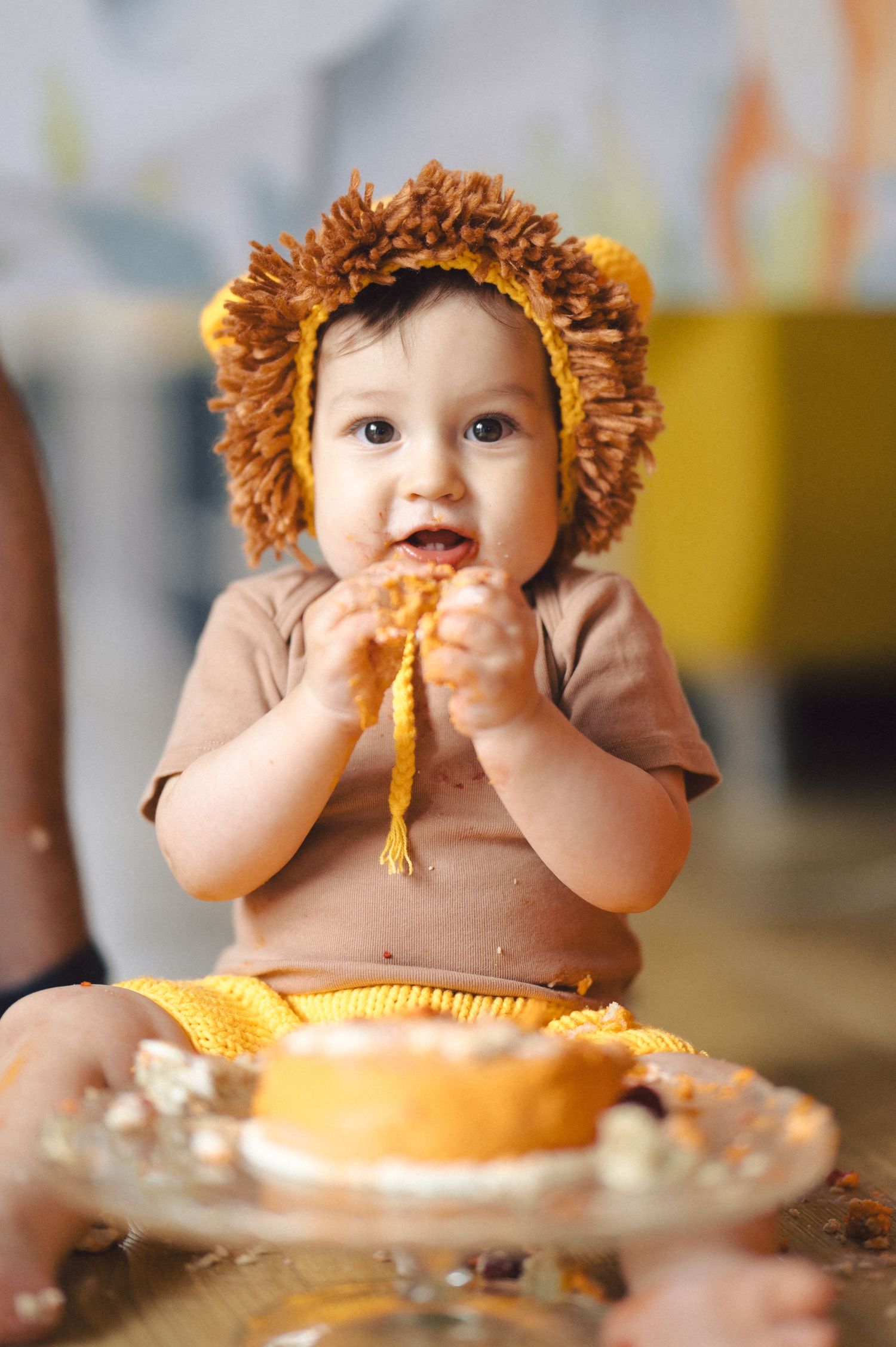 A baby wearing a lion costume sits at a table eating messy food with a plate in front.
