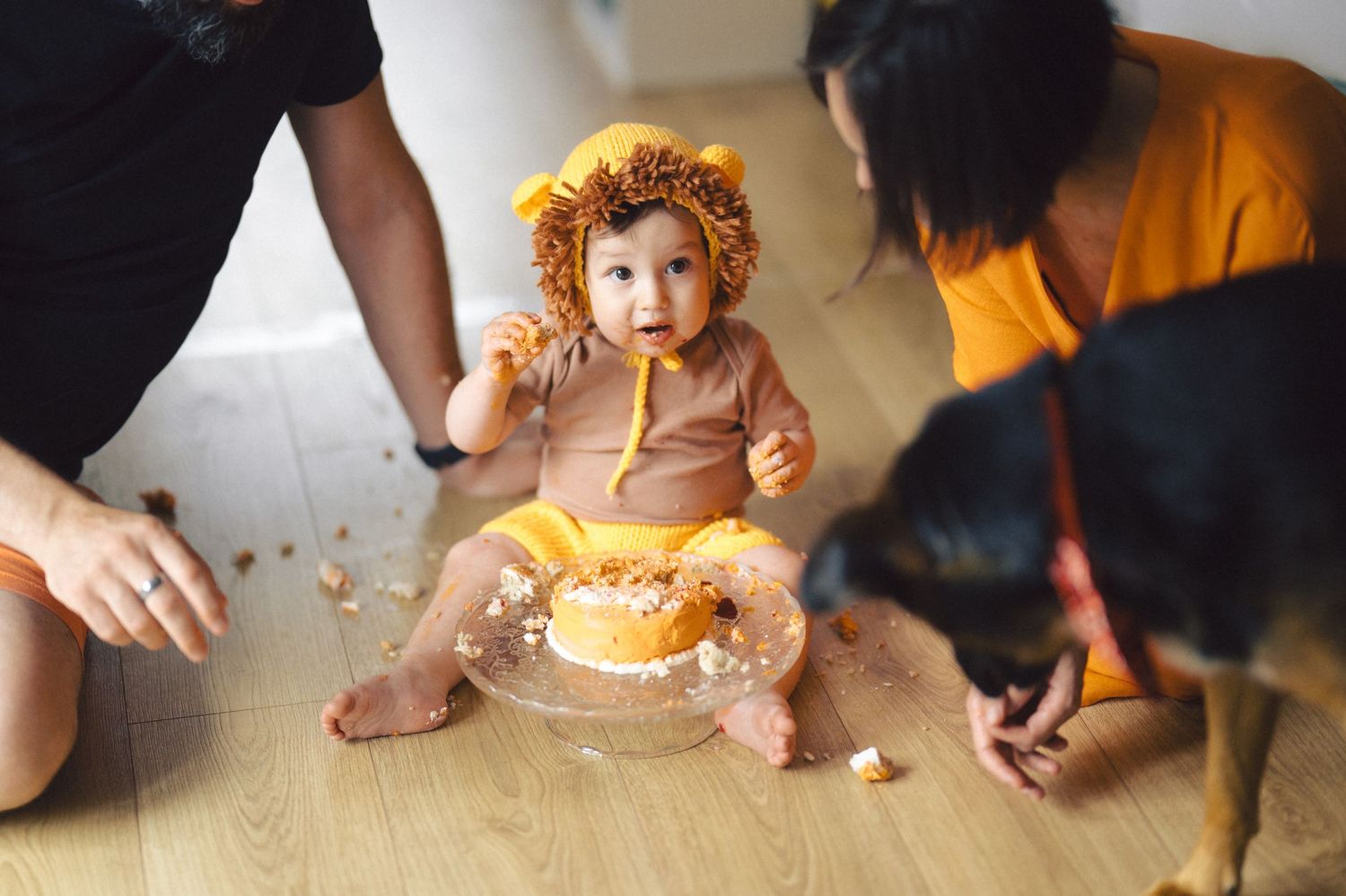 A small figure in a lion costume sits on floor with cake smash celebrating first birthday while family and dog watch nearby.