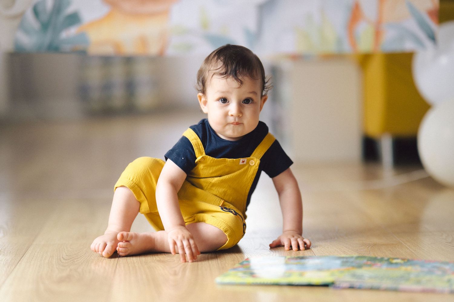 Young child in a yellow romper sits on hardwood floor looking at a book by themselves.