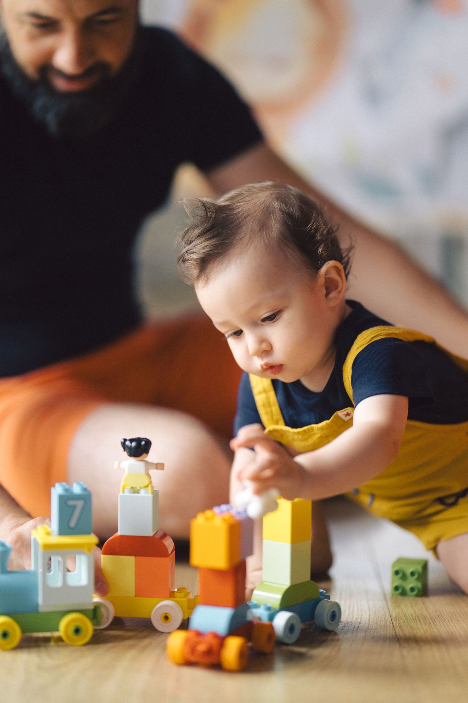 A toddler in a yellow outfit plays with a colorful wooden toy train set while someone watches from behind.