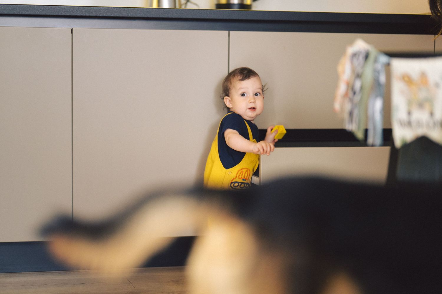 Baby in yellow bib stands with arms outstretched while practicing walking on hardwood floor.