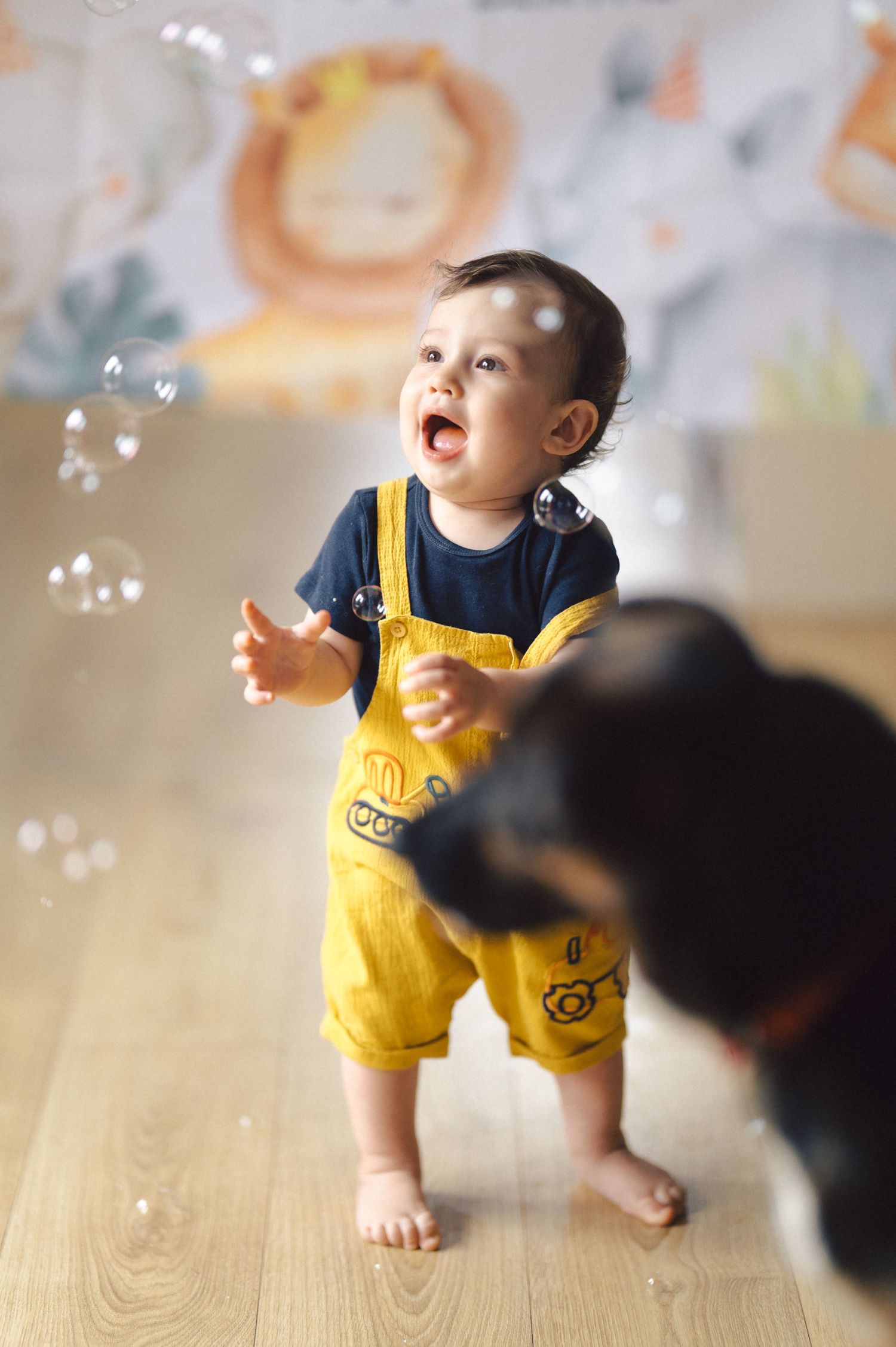 Toddler in yellow overalls stands barefoot on wooden floor playing with floating bubbles while looking up with wonder.
