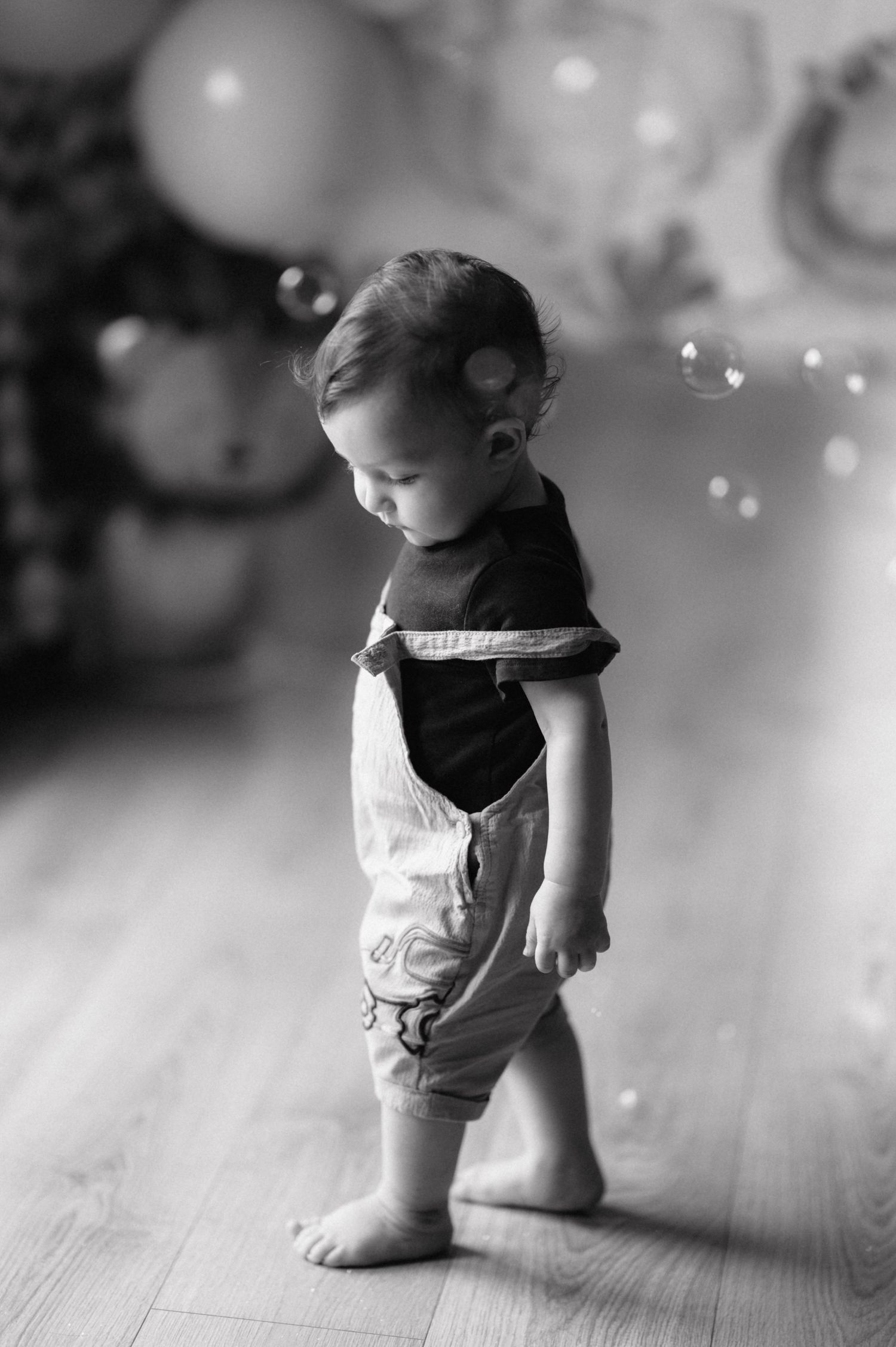 A toddler in overalls stands barefoot on wooden flooring while light bokeh sparkles in the black and white photo.
