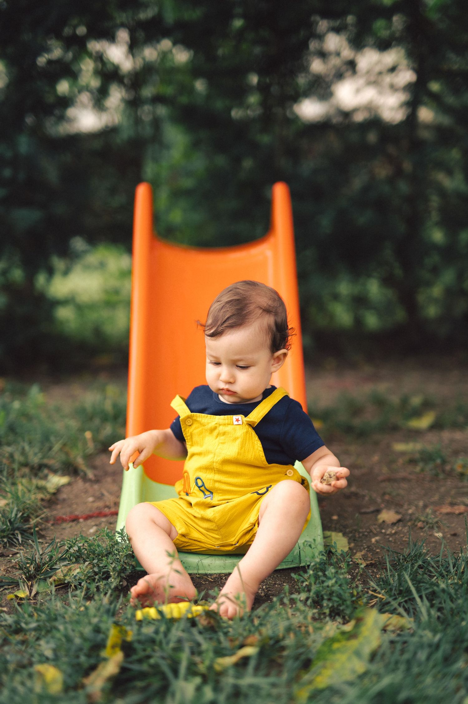 A small child in a yellow romper sits at the bottom of an orange playground slide in a grassy outdoor setting.