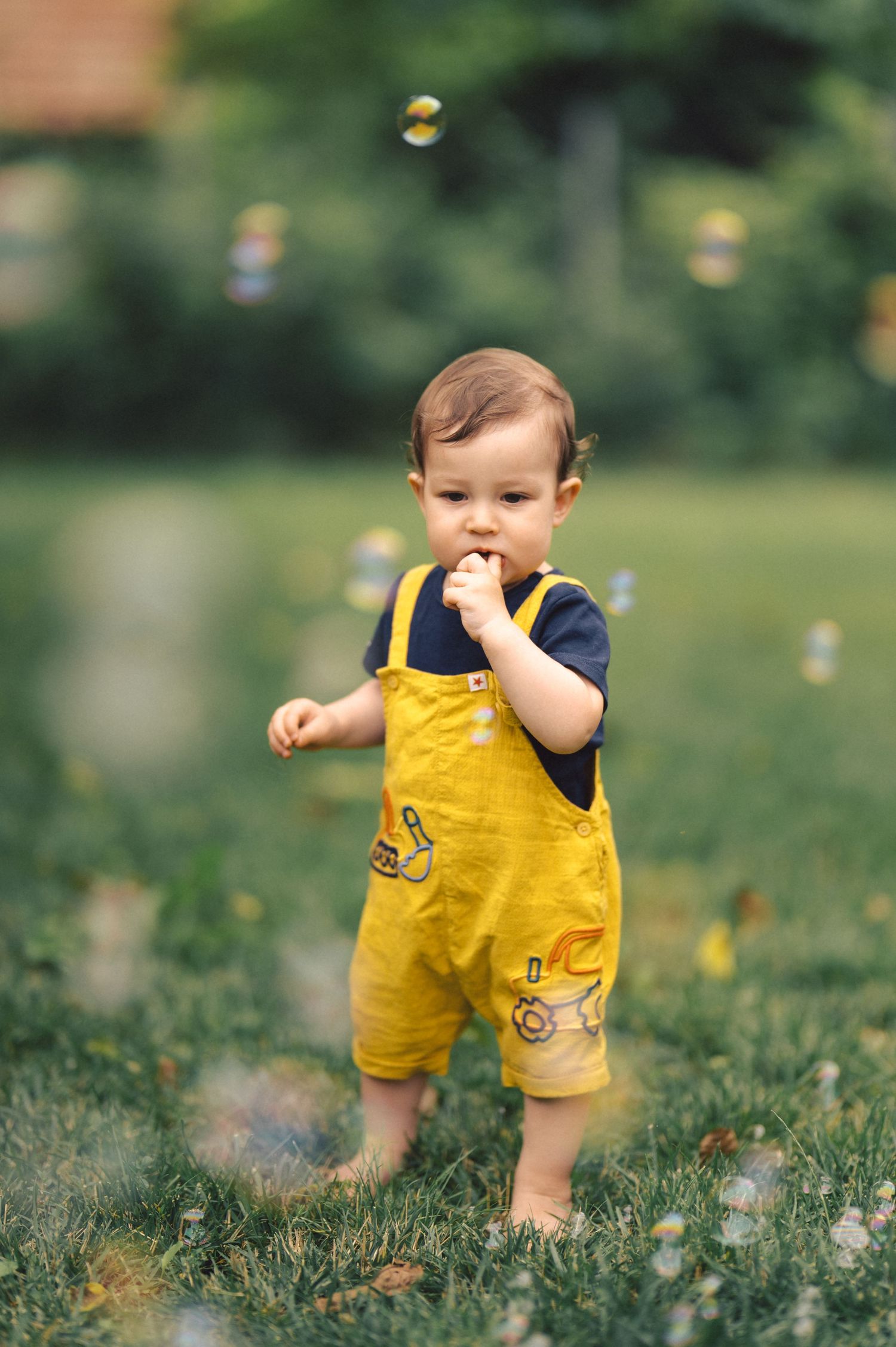 A small child in yellow overalls stands in a grassy field with soap bubbles floating in the air around them.