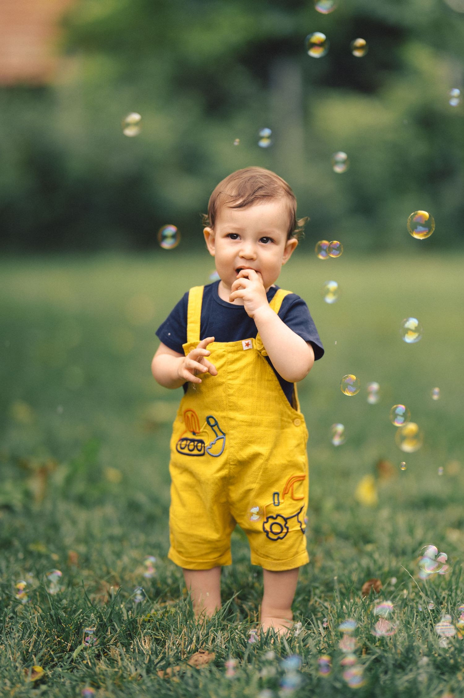 A child wearing yellow shorts plays with soap bubbles floating in a lush green outdoor setting.