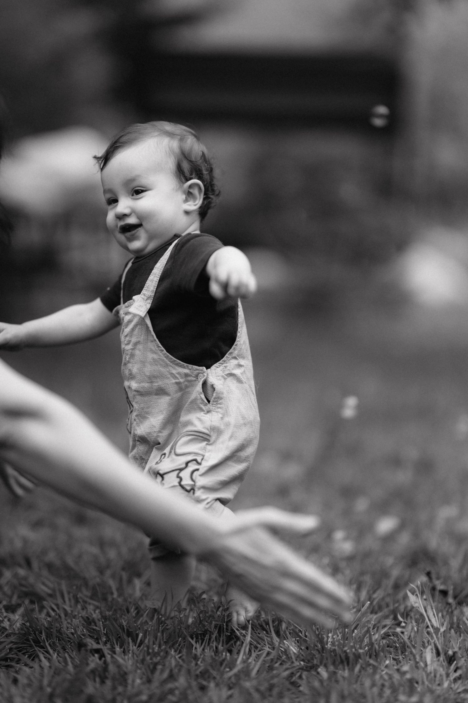 Black and white photo of a joyful toddler wearing denim overalls playing outdoors on a sunny day.