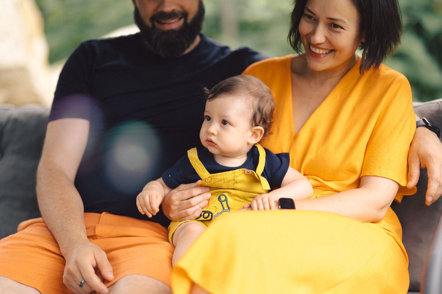 A baby wearing yellow sits between two adults in matching yellow outfits while relaxing outdoors.
