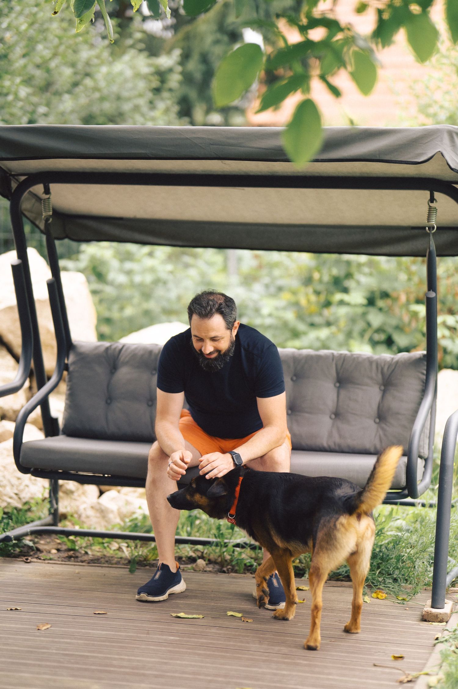 Someone pets a German Shepherd mix dog while sitting on an outdoor porch swing on a wooden deck.