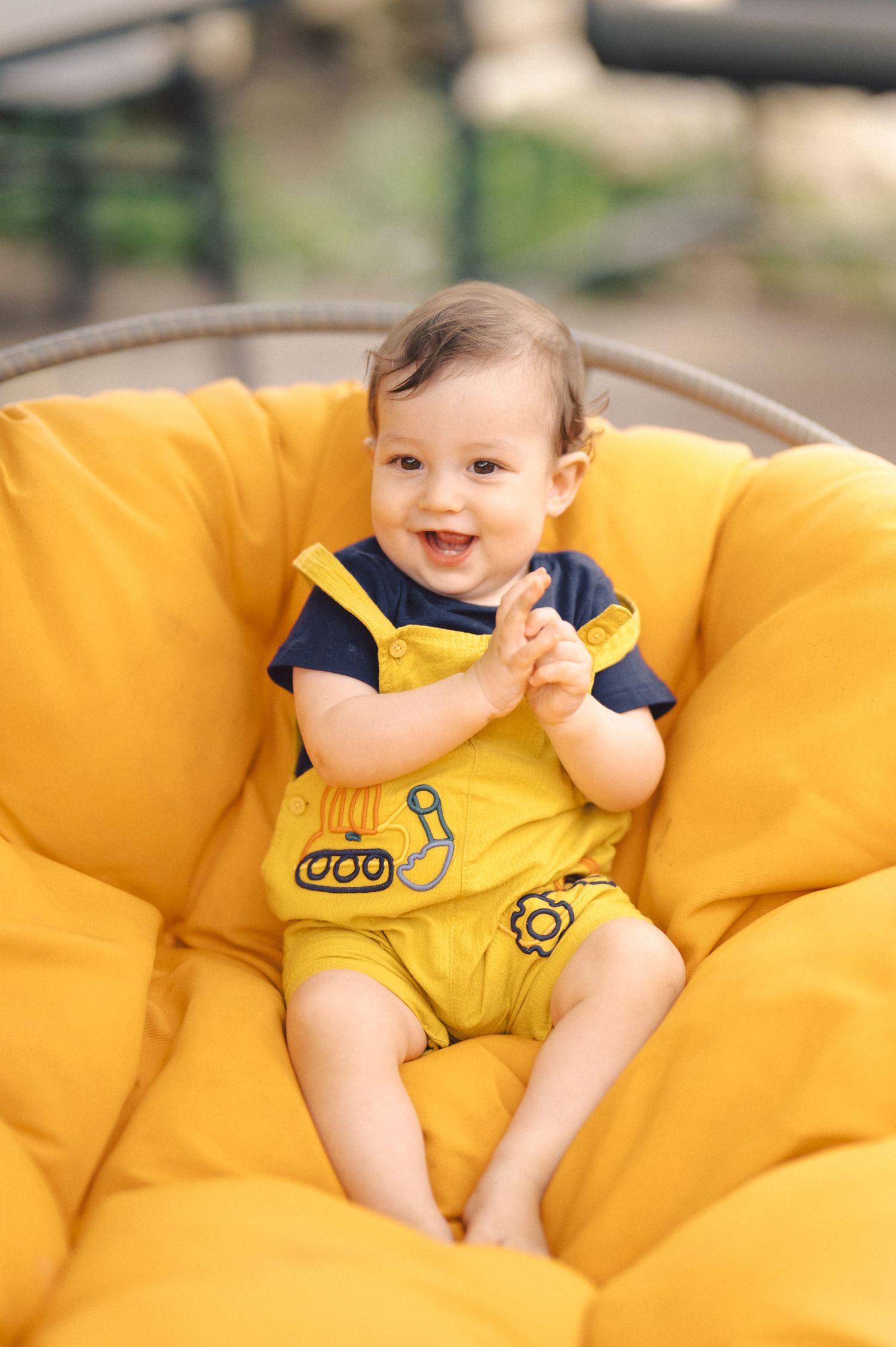 A happy baby wearing yellow overalls with construction designs sits in a plush yellow chair.