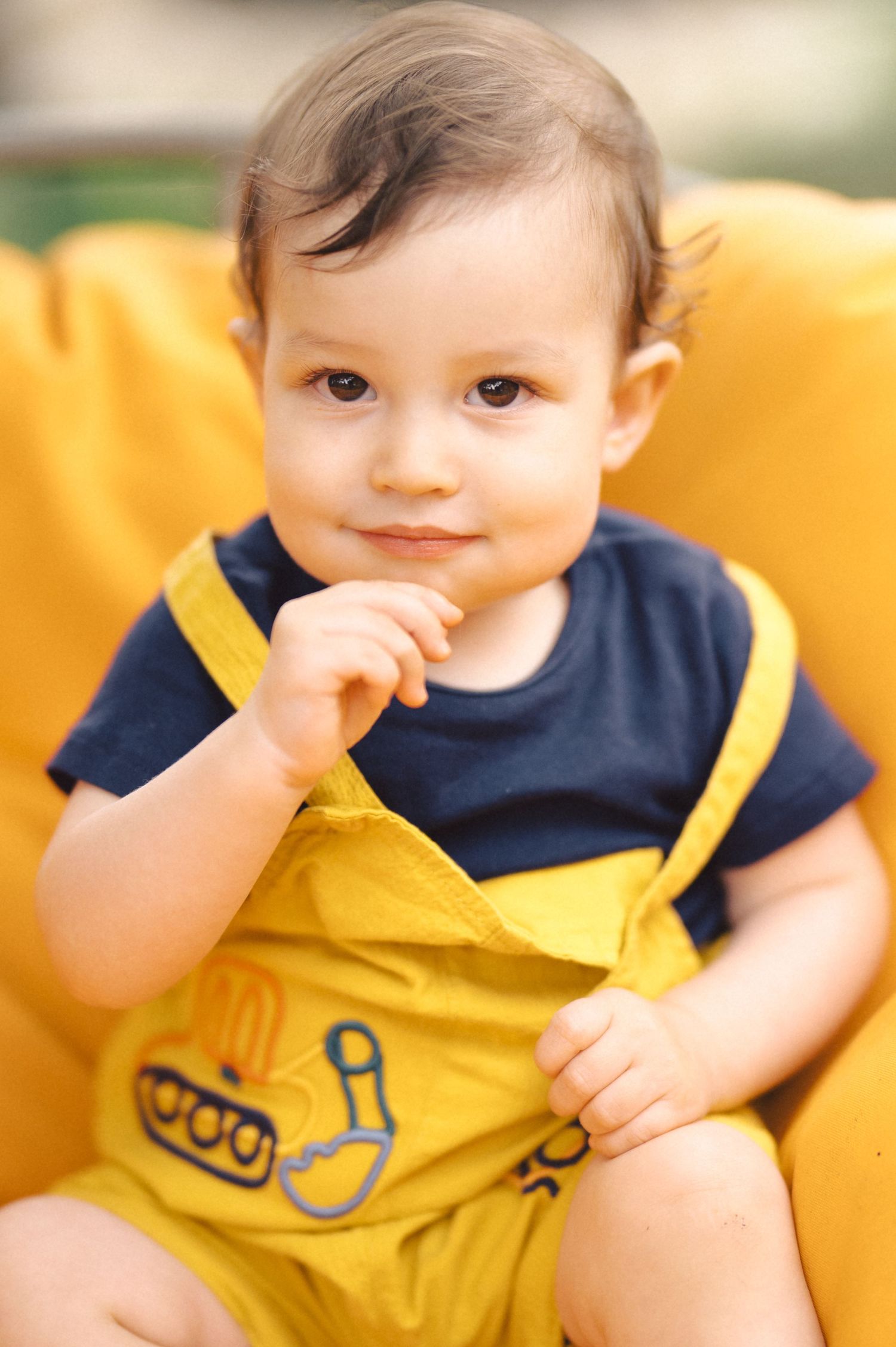 Baby wearing navy and yellow outfit brings fingers to mouth while sitting against yellow background.