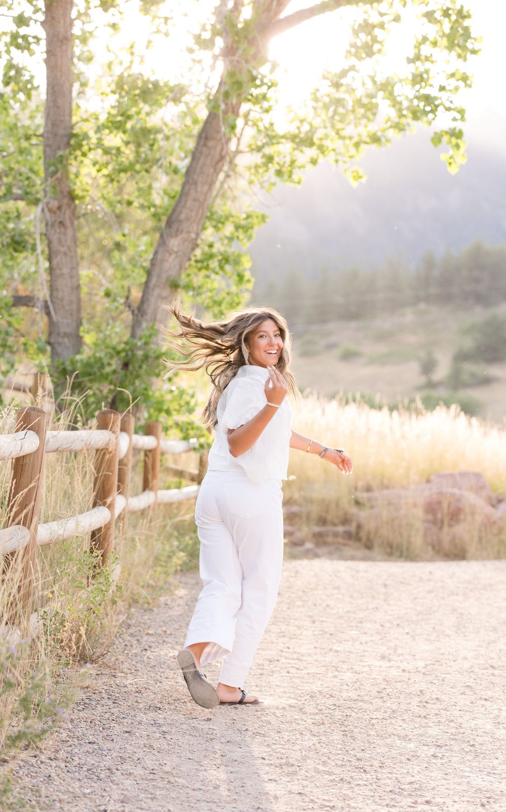 Playful outdoor portrait with flowing hair in all-white outfit along sunlit mountain trail.