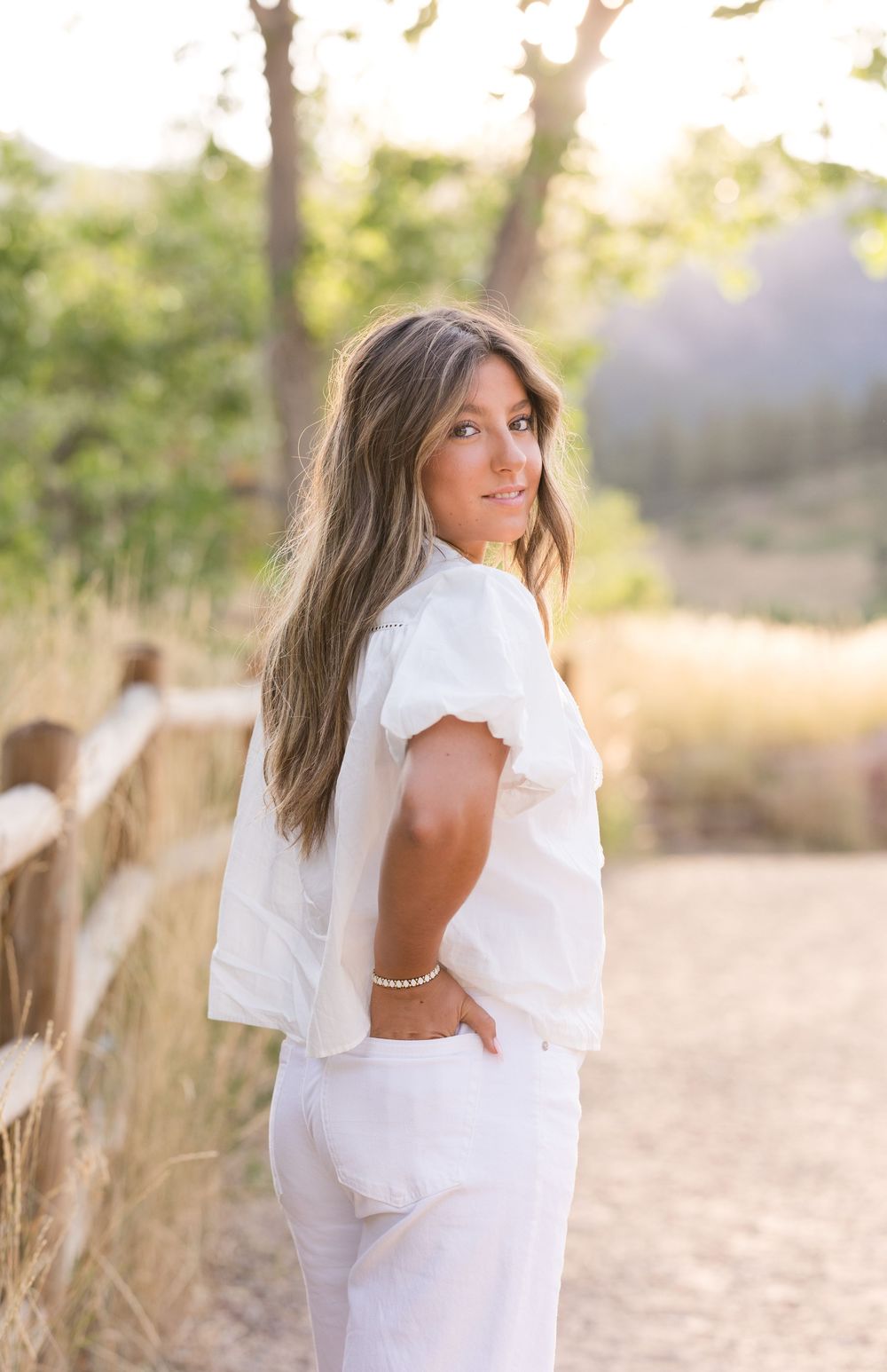 A person in an all-white outfit poses on a dirt path during golden hour with trees and sunlit grass in the background.