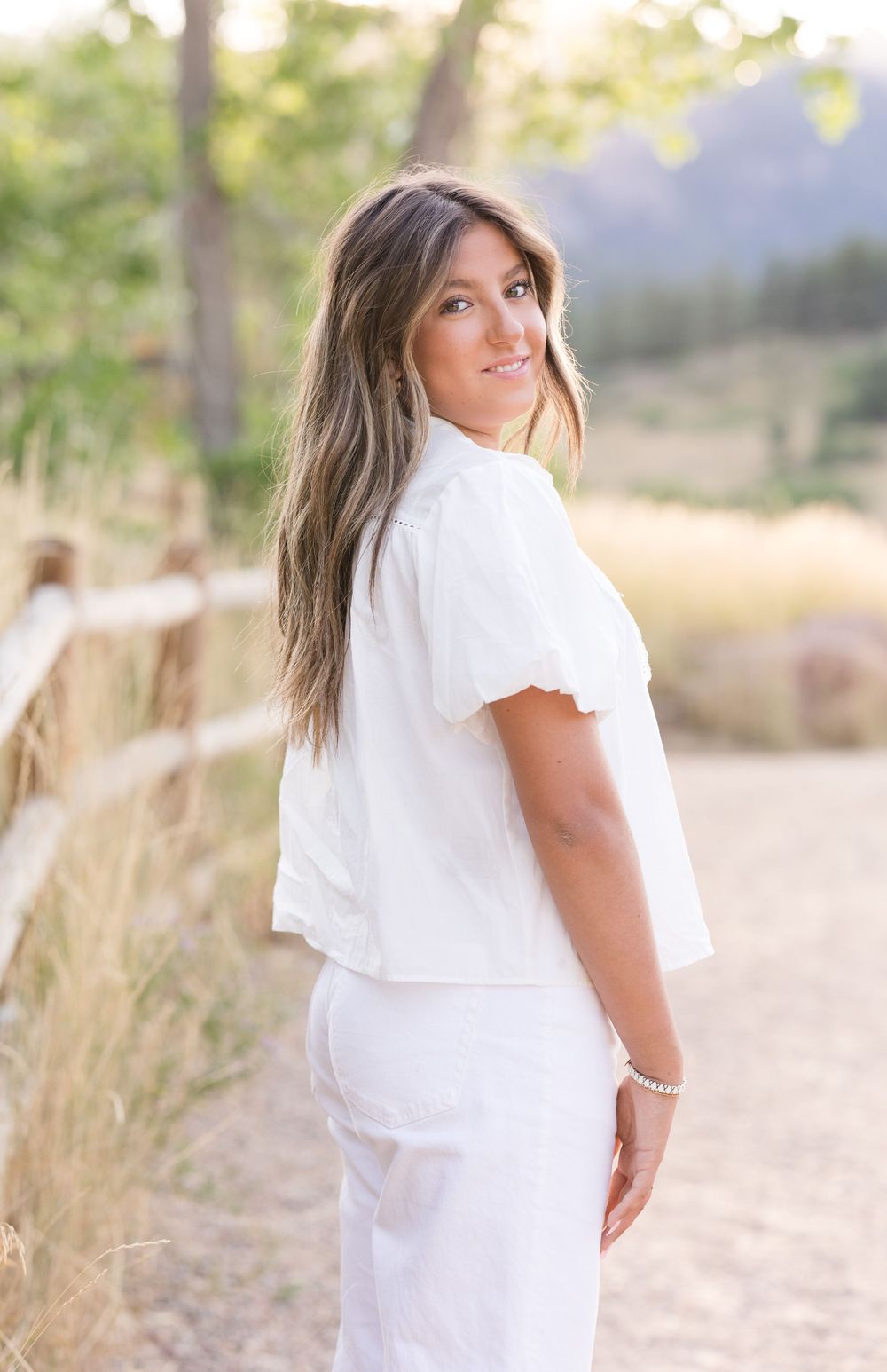 A subject in a white dress poses in a sunlit field with trees creating a dreamy outdoor portrait session effect.
