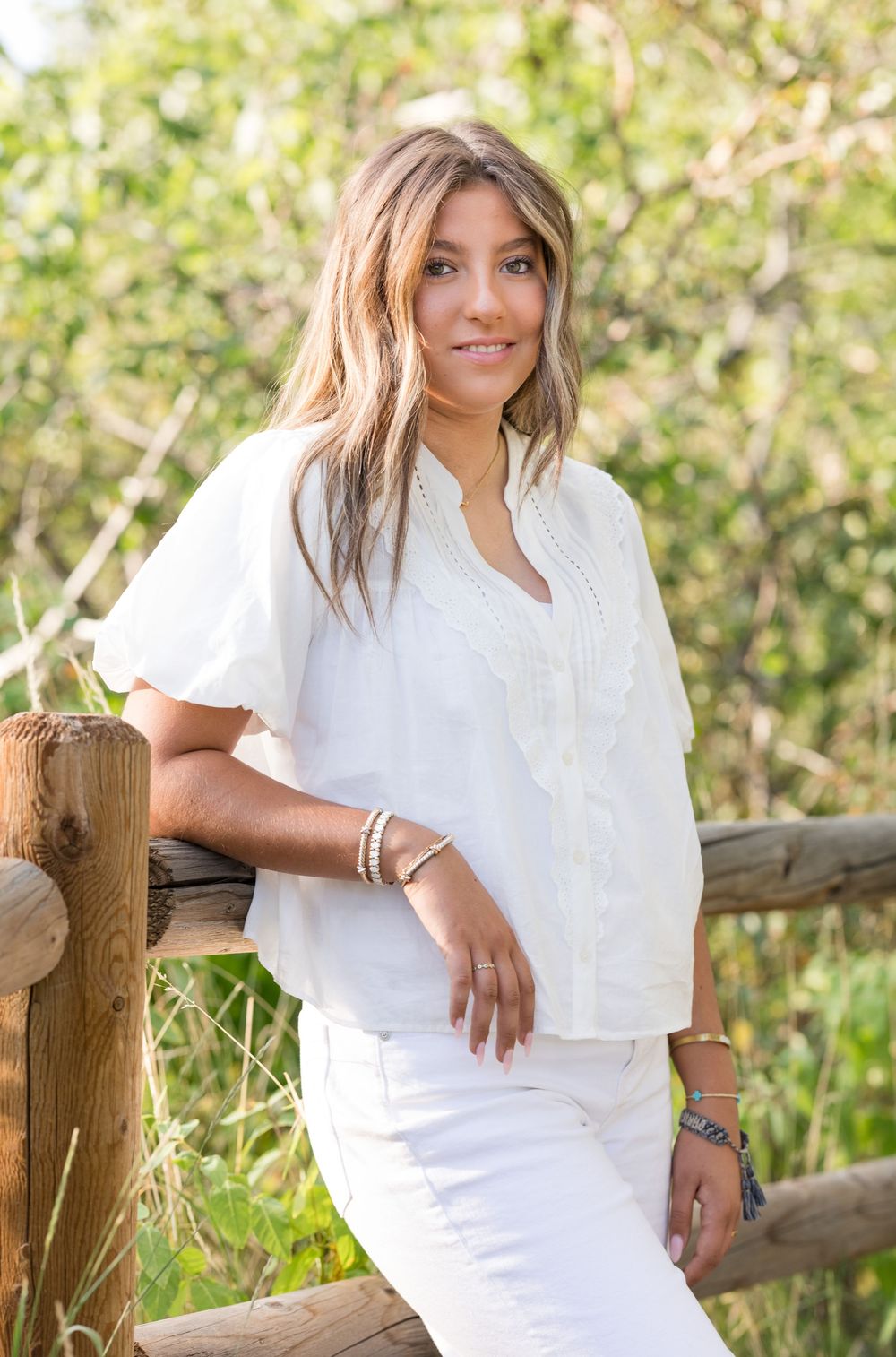 A person in a white dress poses along a rustic wooden fence in an outdoor setting with greenery in the background.