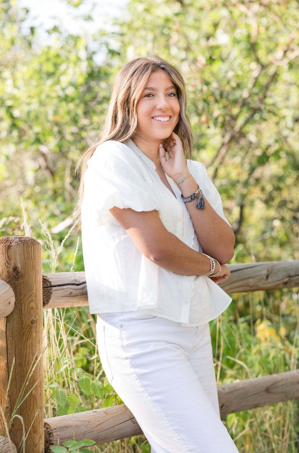 A person in an all-white outfit poses casually against a rustic wooden fence surrounded by summer greenery.