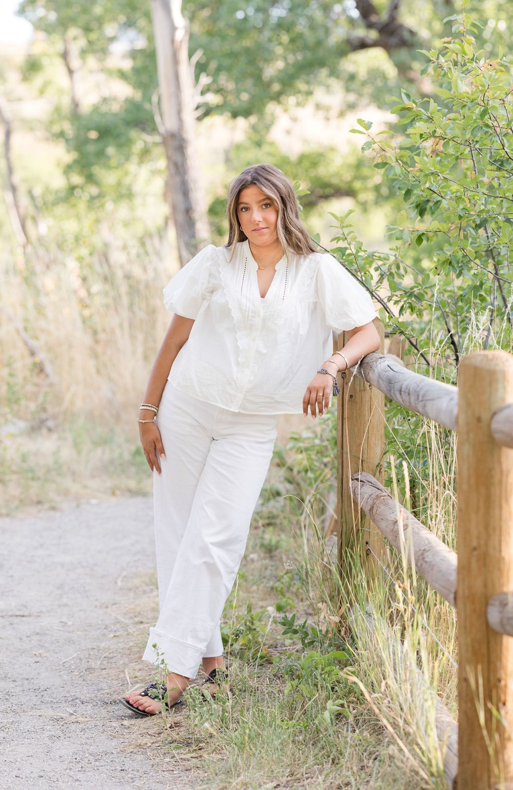 Woman in flowing white outfit poses along rustic wooden fence on dirt path surrounded by natural foliage.