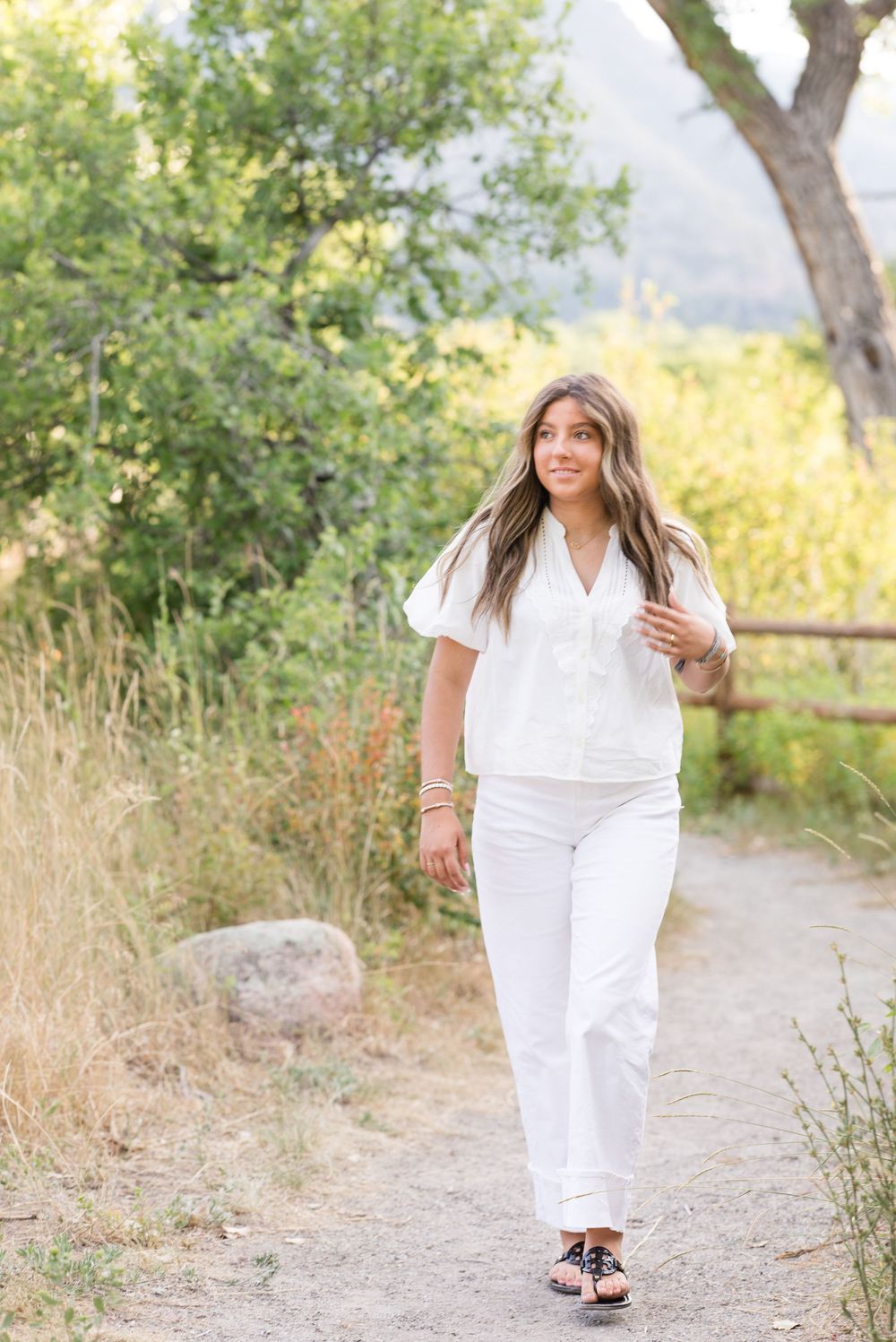 Woman in all-white summer outfit walking along nature trail with trees and mountains in background.