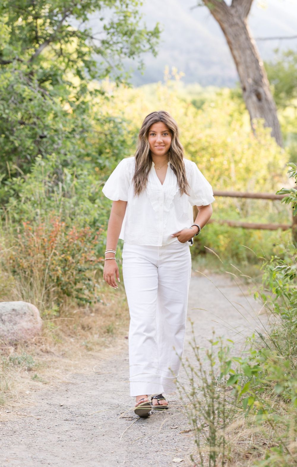 Portrait on dirt path wearing white summer ensemble against scenic mountain backdrop.