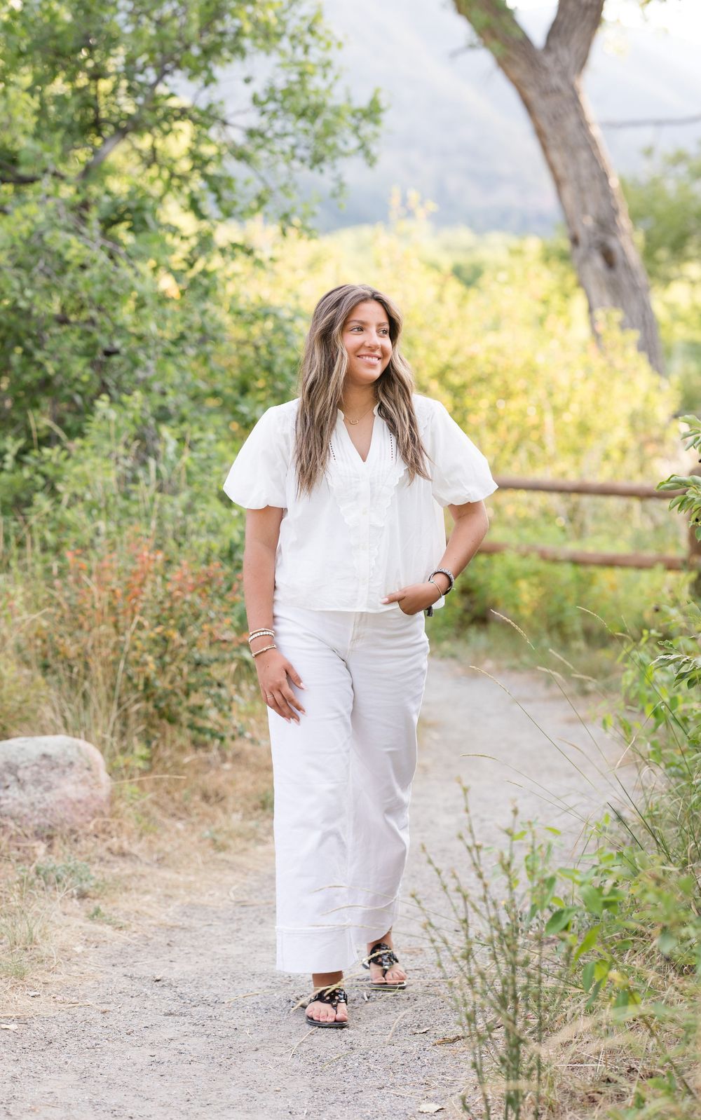 Person in an all-white outfit walks along a nature path surrounded by trees and greenery on a sunny day.
