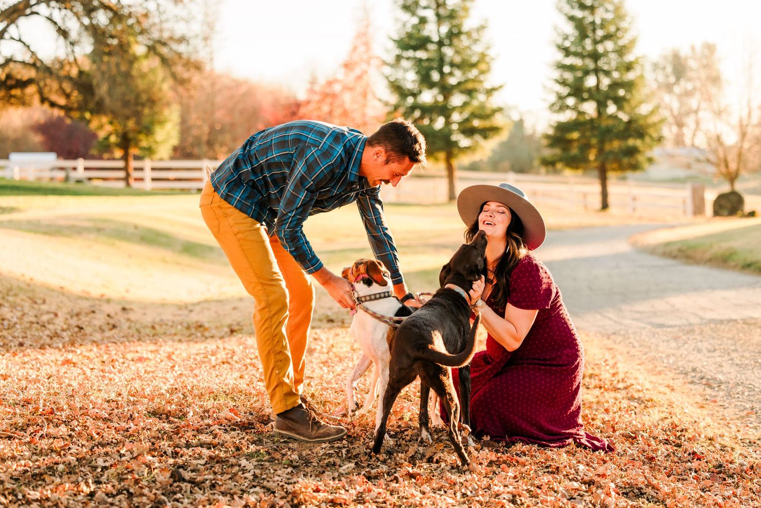 A couple in autumn attire pets their black dog on a path covered with fallen leaves during golden hour.