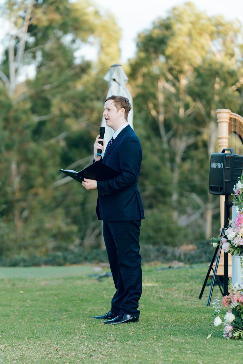 Wedding officiant speaking at outdoor ceremony with floral arrangements.