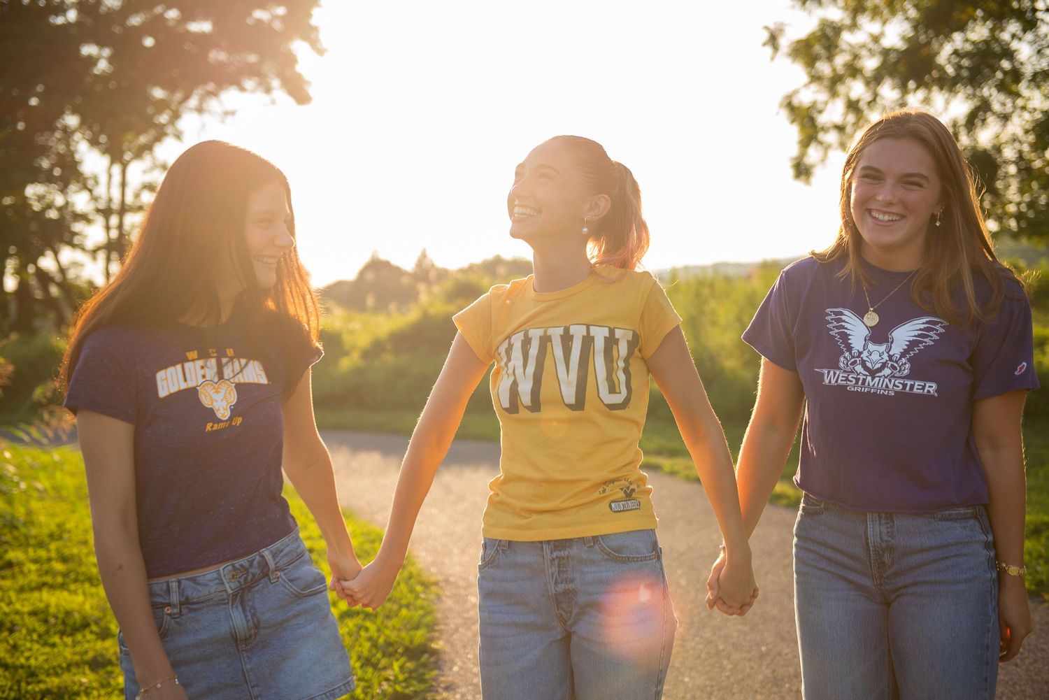 Three friends wearing t-shirts and jeans hold hands while walking together on a path during golden hour sunset.
