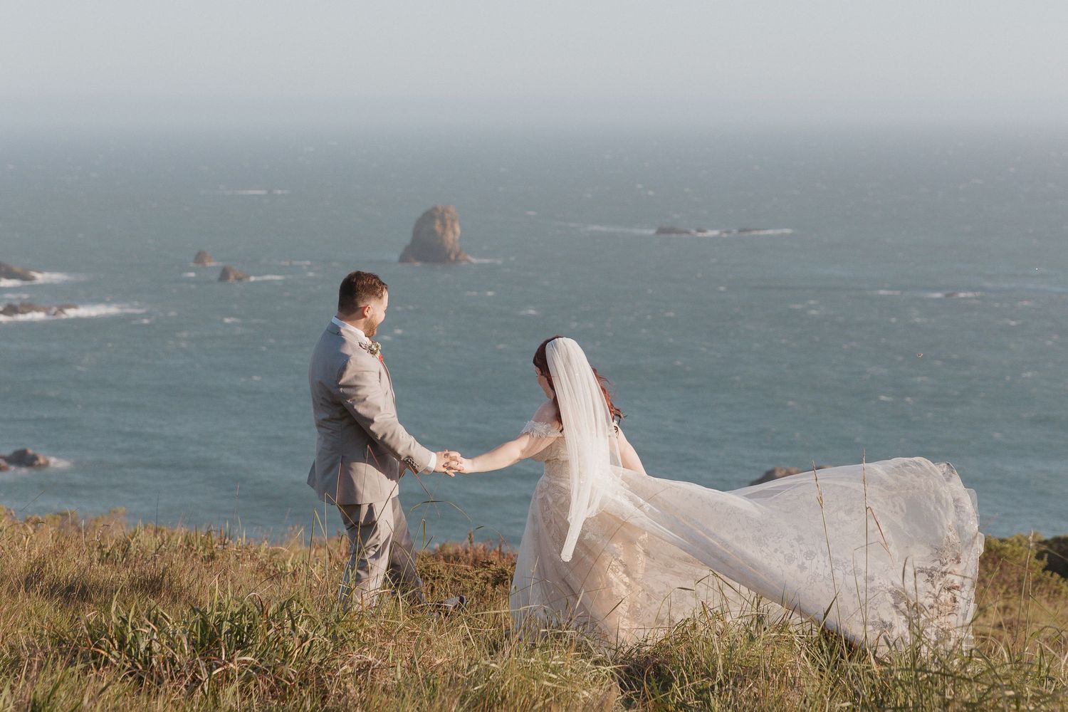 Couple in wedding attire walking along dramatic coastal cliffs overlooking the ocean.