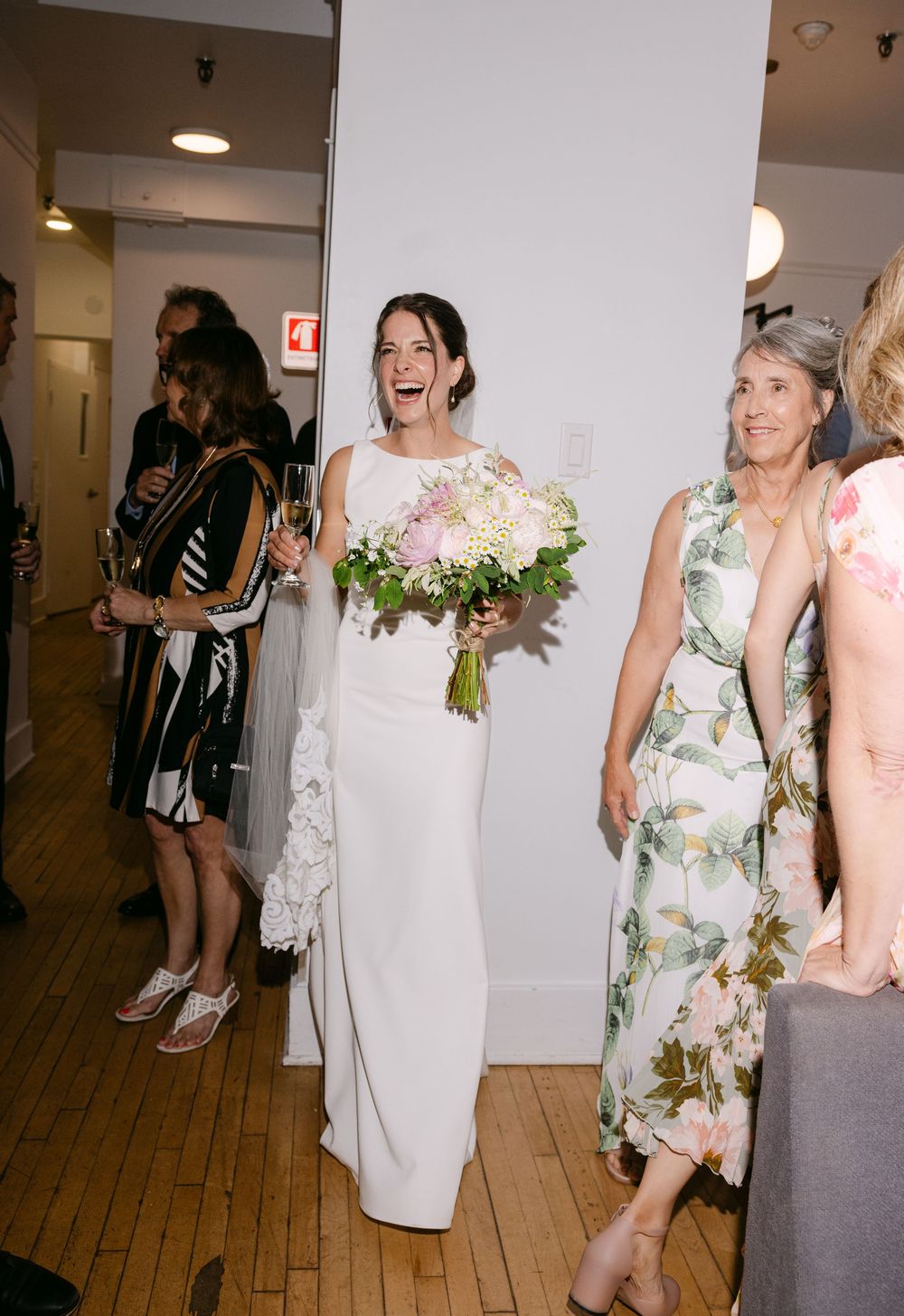 Wedding guests share joyful moments and laughter in an indoor reception venue with white walls and wooden floors.