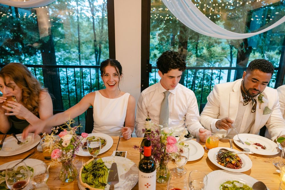 Wedding reception guests enjoy dinner at a decorated table with floral centerpieces and white drapery in the background.