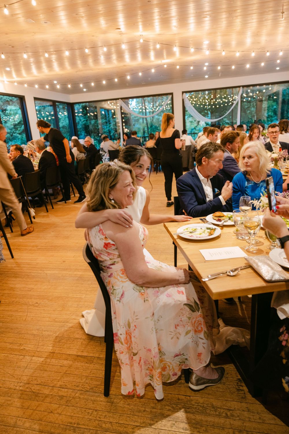Wedding reception guests enjoy dinner and conversation under warm string lights in a rustic venue with large windows.
