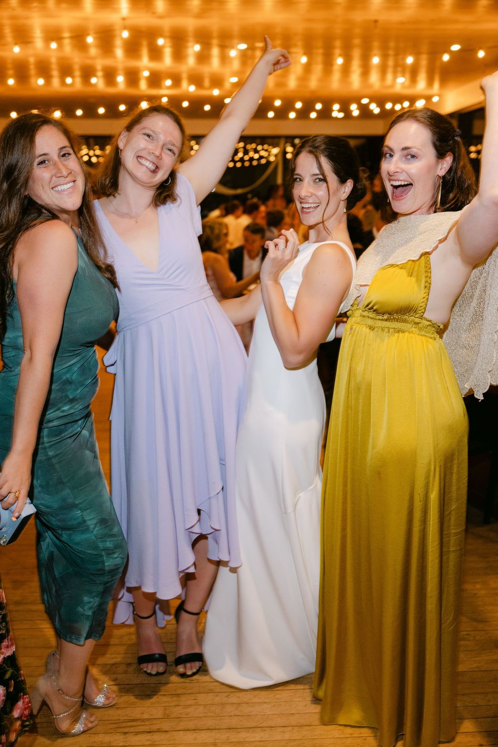 Party guests dancing and celebrating on a wooden dance floor under glowing string lights in green and yellow dresses.