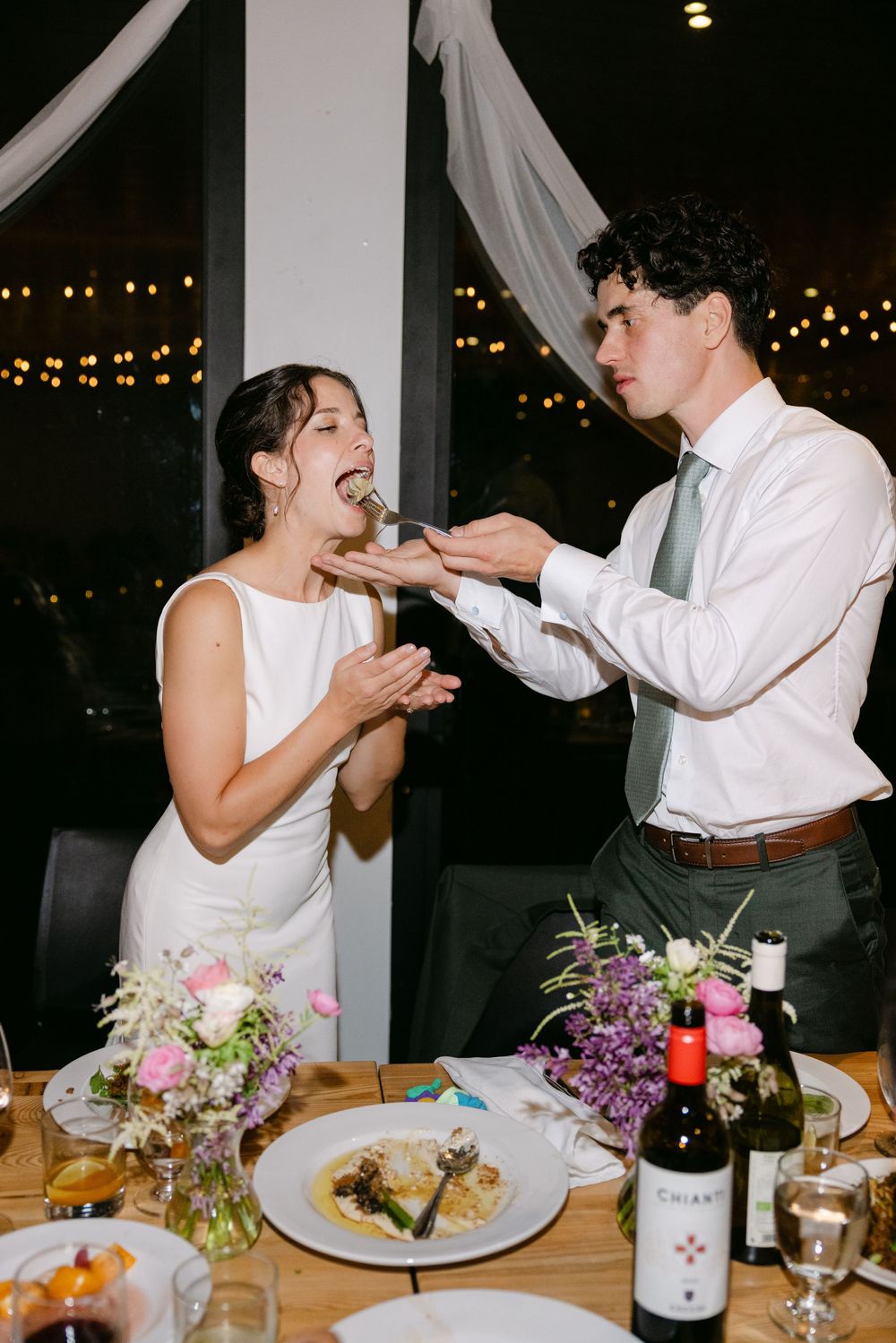 A couple shares a playful cake-feeding moment during their wedding reception at a candlelit dinner table.
