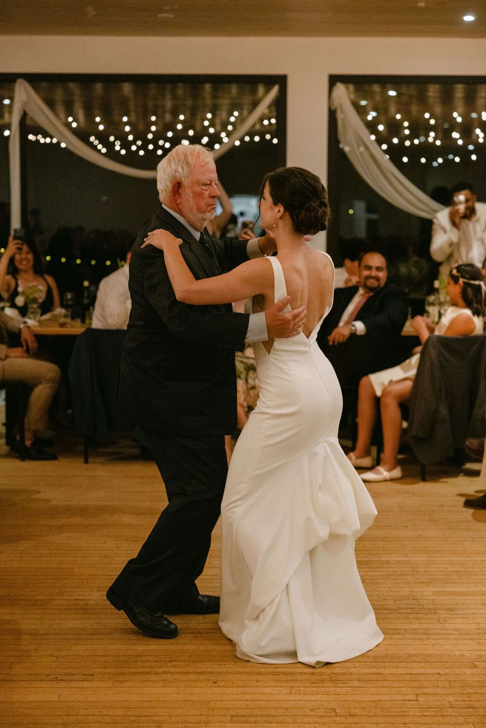 A bride and elderly guest dance on a wooden floor beneath twinkling lights at a wedding reception.