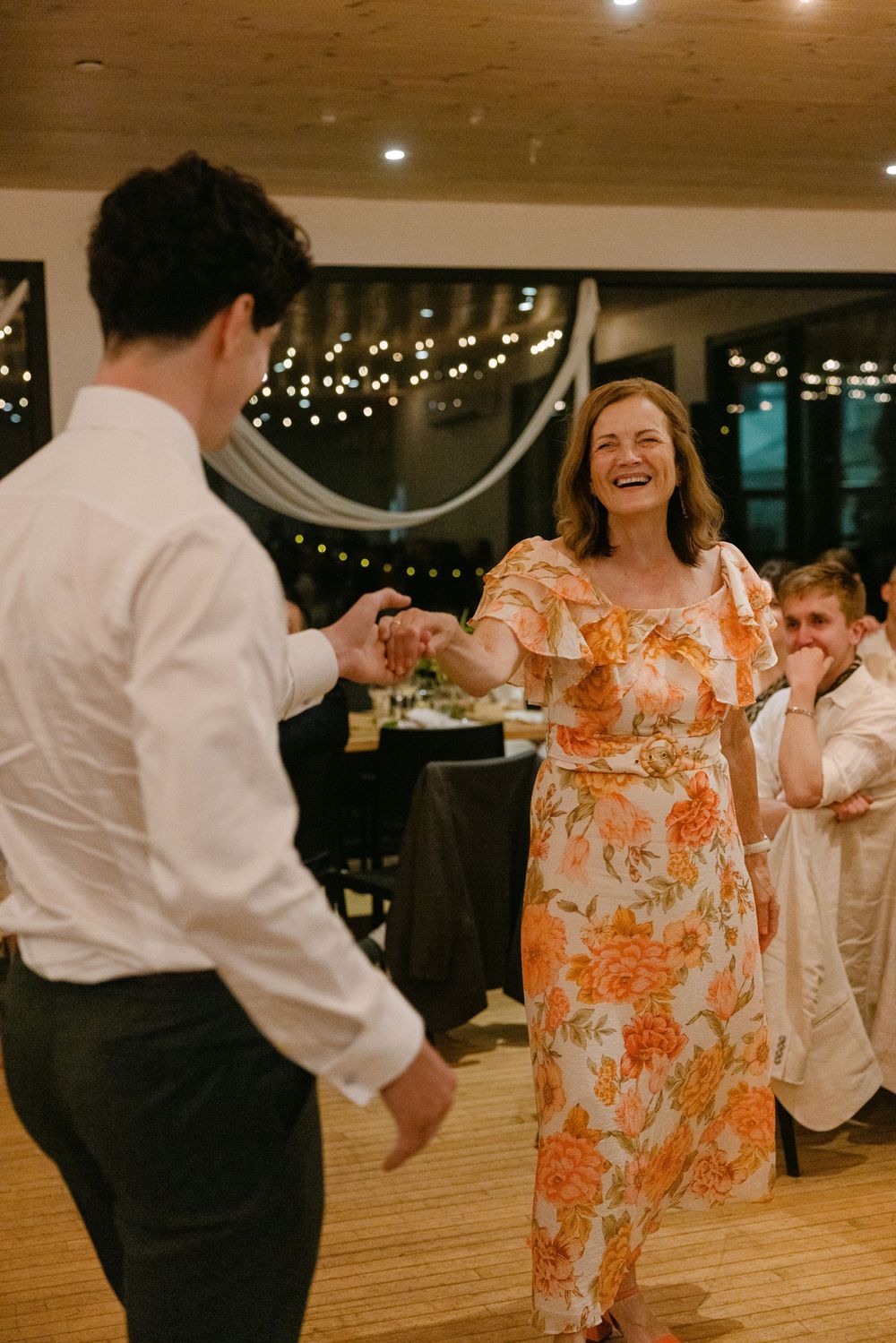 People dance and laugh together at an indoor reception venue decorated with twinkle lights.