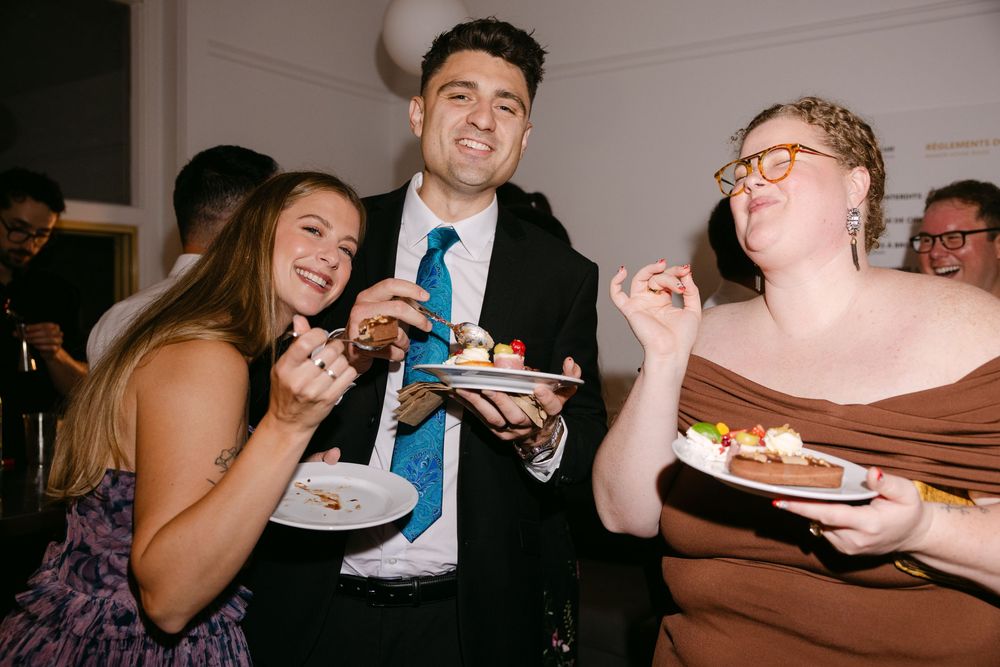 Three party guests enjoying plates of dessert while laughing and socializing at an indoor gathering.