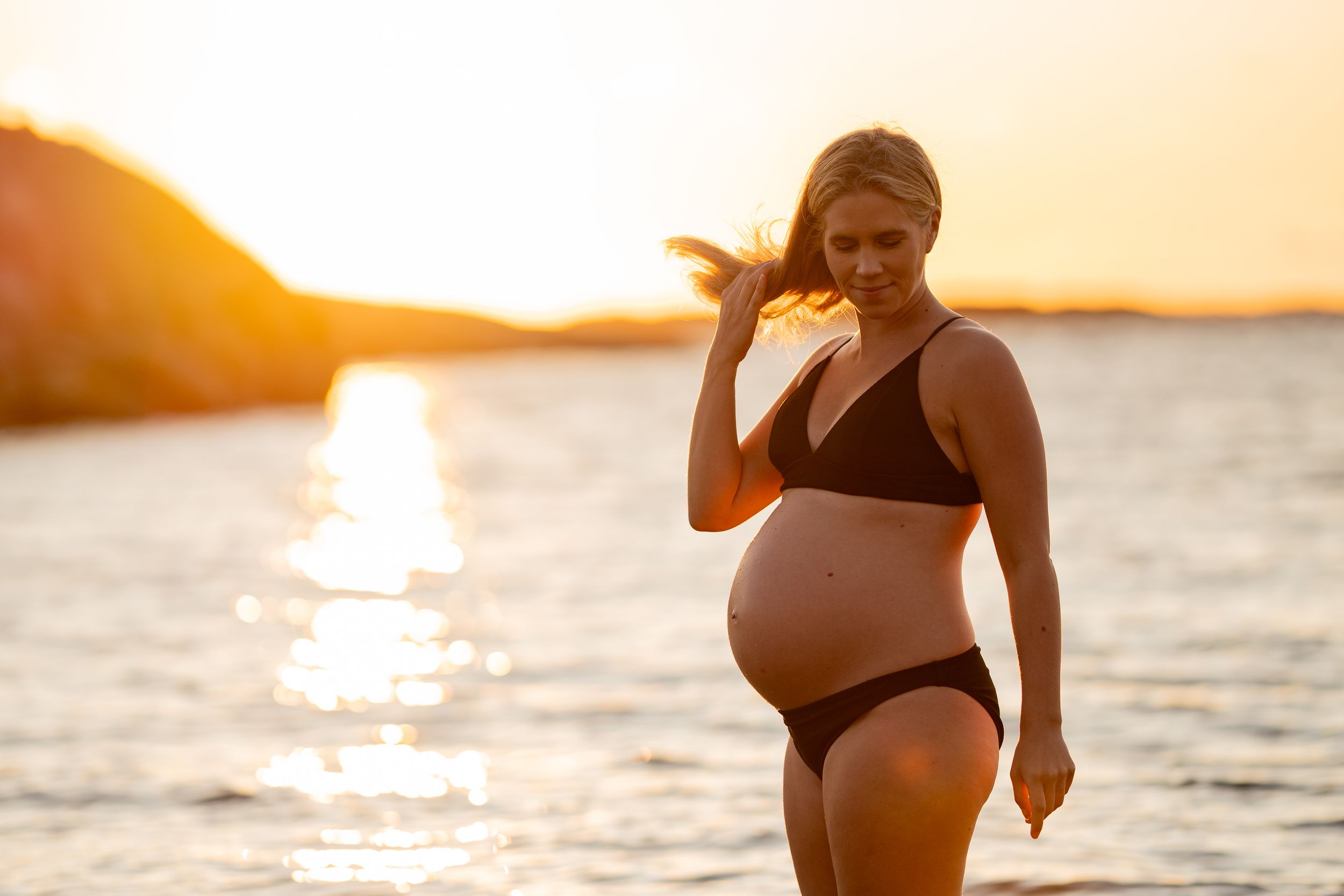En blond gravid kvinna i svart bikini står i havet och håller i sitt hår. I bakgrunden syns solen gå ned i horisonten.