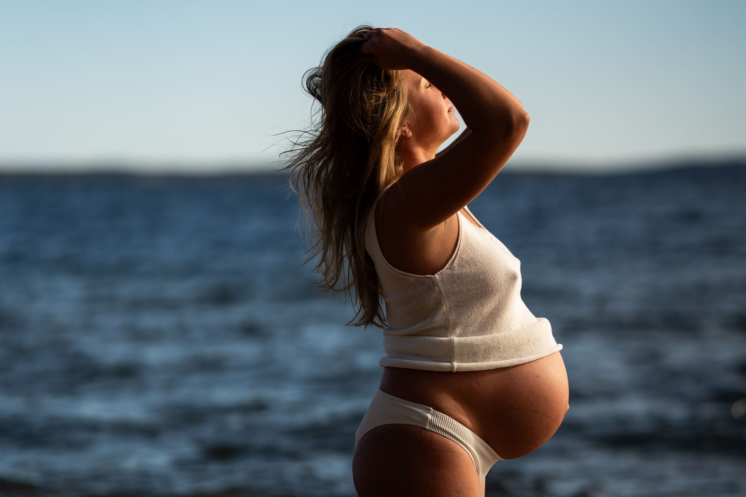 En gravid kvinna med vitt linne och vit bikini står i profil framför havet. Hon blundar mot solen och håller sina armar över huvudet.